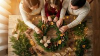 Family making Christmas wreath together at wooden table with pine branches, cinnamon, oranges and festive decorations in warm cozy home atmosphere.