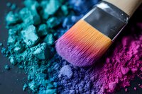 Professional makeup brush with vibrant rainbow bristles surrounded by colorful cosmetic powder pigments in blue, purple, and pink tones for beauty applications.