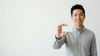Young Asian male professional wearing light gray button-up shirt holding blank white business card against neutral background for corporate branding and contact information display.