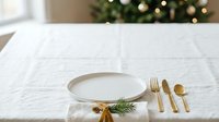 Empty white plate with gold cutlery and evergreen sprigs on white linen tablecloth, festive holiday table setting for dining and celebration events.