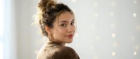Young woman with messy bun hairstyle wearing cozy knit sweater smiling over shoulder in bright interior with bokeh lights for lifestyle content.