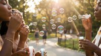 Two young women blowing soap bubbles in sunny park during golden hour creating magical floating spheres in warm sunlight for friendship and joy concept.