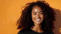 Young African American woman with curly hair smiling against orange wall background, radiating happiness and confidence in warm sunlight.