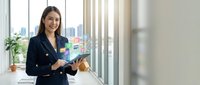 Smiling young Asian businesswoman in navy suit holding tablet with colorful app icons floating above screen in modern office.