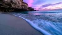 Dramatic sunset seascape with purple sky over rocky coastline and foaming waves washing onto sandy beach creating peaceful coastal atmosphere.