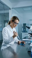 Female scientist in white lab coat and glasses conducting research with petri dish and microscope in modern laboratory setting.