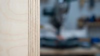 Plywood edge detail with blurred workshop background showing woodworking tools and equipment for construction and carpentry projects.