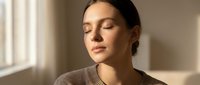 Young woman with dark hair and olive complexion, wearing a neutral beige sweater, eyes closed in peaceful expression, soft natural window lighting, calm and serene mood for wellness and mindfulness content.