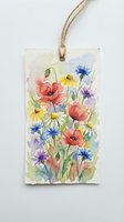 Watercolor wildflower arrangement featuring red poppies, blue cornflowers, yellow daisies, and purple blooms with green stems on textured paper tag with twine.