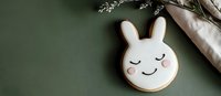Cute white bunny sugar cookie with pink cheeks and closed eyes on dark green background. Homemade Easter treat with minimal decoration style.
