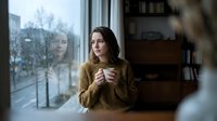 Young woman with long dark hair and fair complexion, wearing a mustard yellow ribbed sweater, holding a warm beverage while gazing thoughtfully out a window on a winter day. Suitable for lifestyle, wellness, and seasonal content.