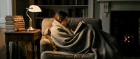 Young woman of mixed ethnicity relaxing on armchair wrapped in cozy blanket, reading book by warm lamp light in comfortable home library setting with fireplace.