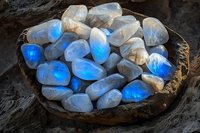 Glowing moonstone crystals in rustic wooden bowl emit ethereal blue light against dark background, creating mystical mineral display for spiritual and wellness concepts.