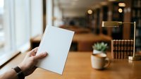Caucasian hand holding blank white card in modern office workspace with wooden desk, blurred background, and natural lighting for branding and marketing mockups.