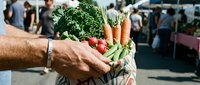 Man holding fresh vegetables at farmers market with produce stand and shoppers in background.