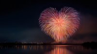 Heart-shaped fireworks display illuminating night sky over calm water with city skyline silhouette in background, perfect for celebration themes.