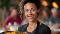 Young African American female server in restaurant holding plate of fresh salad, wearing black uniform and natural curly hair, genuine smile at upscale dining venue.