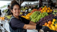 Hispanic female produce worker arranging fresh fruits at farmers market stall, displaying oranges and grapes while wearing black uniform and gloves.