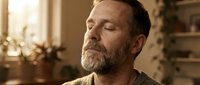 Middle-aged man with gray beard and brown hair in contemplative pose, wearing neutral toned shirt in warm home interior with natural lighting and blurred background.