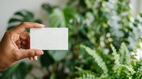 Person with medium skin tone holding blank white business card against lush green foliage background, suitable for branding, corporate identity, and eco-friendly business concepts.