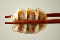 Asian dumpling held by wooden chopsticks showing translucent wrapper and pleated edges, with chili oil spots on neutral background. Great for food menu design.
