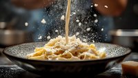 Freshly grated parmesan cheese falling onto steaming fettuccine pasta in dark ceramic bowl with dramatic lighting and motion capture of cheese particles.
