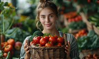 Young Caucasian woman holding wicker basket full of fresh ripe tomatoes in greenhouse, smiling at camera. Natural organic farming and local produce concept.
