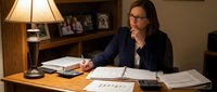 Professional businesswoman with glasses working late at office desk reviewing financial documents and charts under warm lamp light.