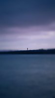Solitary figure standing on distant shoreline at twilight with calm water and purple-pink sky, conveying contemplation and isolation for editorial and lifestyle projects.