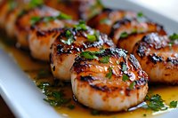 Grilled sea scallops with butter sauce and fresh herbs on white plate, close up view showing caramelized sear marks and garnished with chopped parsley.