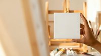 Hand holding blank white card in front of wooden easel with art supplies, suitable for mockups, presentations, creative business concepts, and design templates.