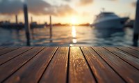 Wooden pier deck at sunset with blurred marina and yacht silhouettes in background, golden sun reflection on calm water creates peaceful nautical atmosphere.