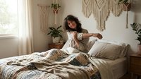 Young woman with dark hair and medium skin tone sitting on unmade bed in boho bedroom with macrame wall hangings and potted plants, wearing casual beige clothing, morning lifestyle scene.