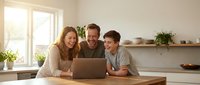 Happy family with teenage son using laptop together in modern kitchen during morning sunlight for online learning or entertainment activities.