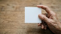 Mature man with weathered skin holding blank white card against wooden surface for mockup and branding purposes.