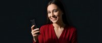 Happy young Caucasian woman in red blazer smiling while using smartphone against dark background for business communication and technology concepts.