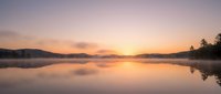 Serene lake at sunrise with golden light reflecting on calm water, misty mountains and evergreen trees silhouetted against pastel sky.