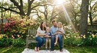 Young Caucasian family with two children sitting on wooden bench in blooming spring garden with sunlight filtering through oak trees.