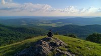 Person sitting on mountain peak overlooking vast green valley and rolling hills under blue summer sky with white clouds, peaceful meditation moment.