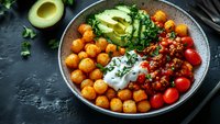 Healthy bowl with crispy chickpeas, ground meat, cherry tomatoes, avocado slices, fresh herbs and sour cream served on dark background for food blogs.