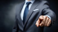 Businessman in elegant gray suit pointing finger forward against dark background, conveying leadership and decision-making in corporate environment.