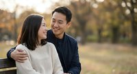 Happy Asian couple embracing on park bench during golden hour sunset, romantic relationship moment outdoors in autumn setting for lifestyle content.