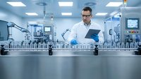 Male scientist in white lab coat and safety glasses reviewing data on tablet while monitoring automated pharmaceutical production line in modern sterile facility.
