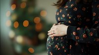 Pregnant woman in floral dress gently holding baby bump against blurred Christmas lights background, creating intimate maternal moment with soft bokeh effect.