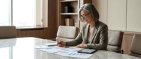 Mature woman with gray hair and glasses wearing business casual attire working at marble desk with laptop, tablet and documents in modern office environment with natural lighting.