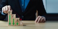 Professional businessman arranging wooden blocks with declining green arrows showing downward trend and decreasing numbers in corporate office setting.