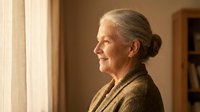 Senior Caucasian woman with gray hair in bun wearing brown cardigan standing by window in warm indoor lighting for lifestyle and aging concepts.