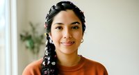 Young woman with long black braided hair adorned with white flower clips, wearing an orange-brown sweater, smiling at camera in bright indoor setting with plants.