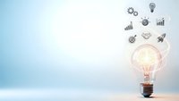 Glowing light bulb with business icons floating above on blue gradient background representing innovation, creativity and strategic planning concepts.