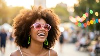 Young African American woman with curly afro hair wearing pink sunglasses and green earrings smiling joyfully at outdoor summer festival with bokeh lights.